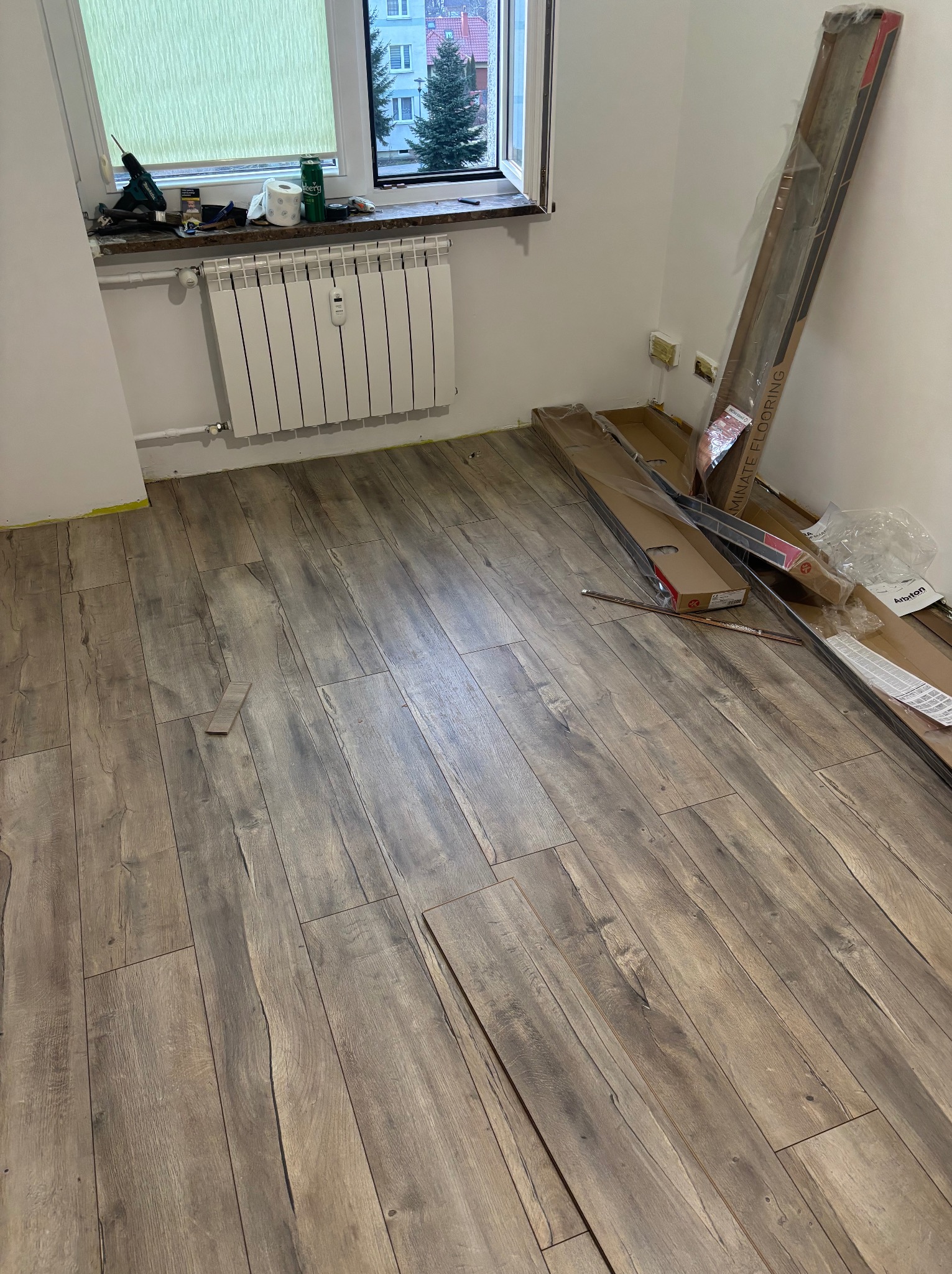 Układanie paneli podłogowych w pokoju. Częściowo ułożona podłoga z paneli w odcieniach brązu, paczki paneli przy ścianie, narzędzia na parapecie okna.