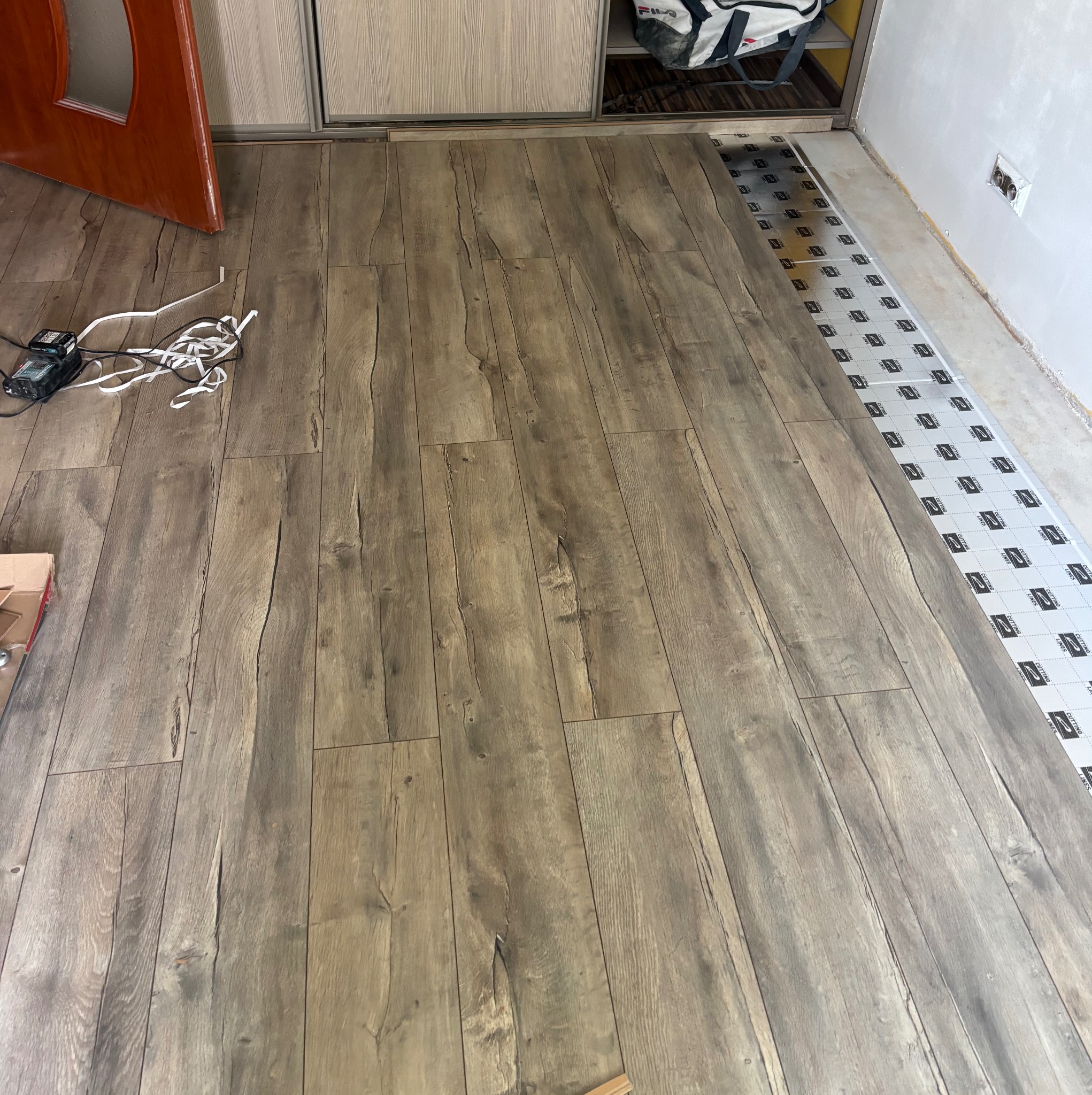 Ułożone panele podłogowe o wzorze drewna, widoczne narzędzia i podkład. Częściowo widoczna ościeżnica drzwi i szafa przesuwna. Podłoże przygotowane pod dalszy montaż.