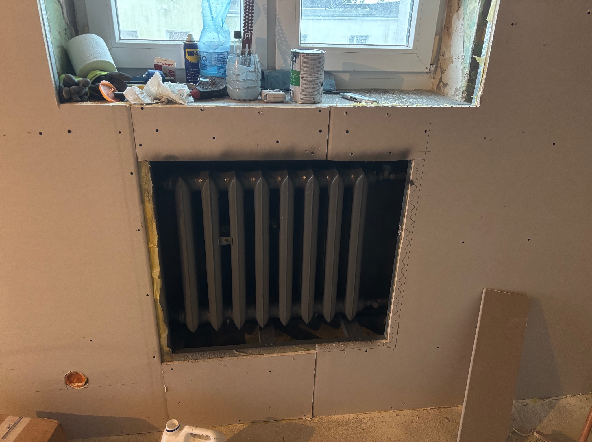 Ściana z płyt karton-gips, otwór na grzejnik w trakcie zabudowy. Widoczne narzędzia i materiały budowlane na parapecie okna. Żółta izolacja w szczelinach.