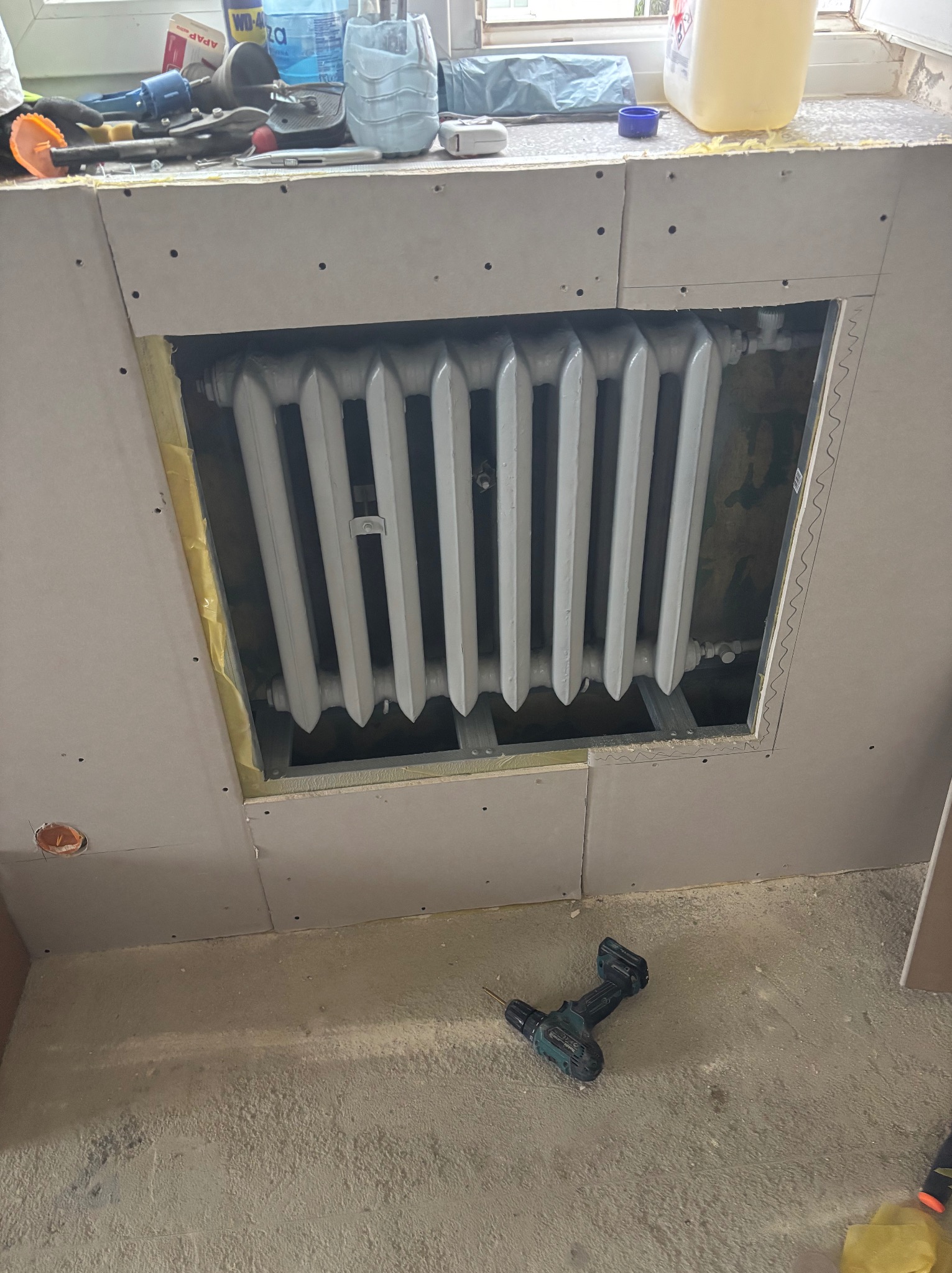 Grzejnik w obudowie z płyty karton-gips, widoczne narzędzia na parapecie i wiertarka na podłodze. Etap prac wykończeniowych, widok z dołu.