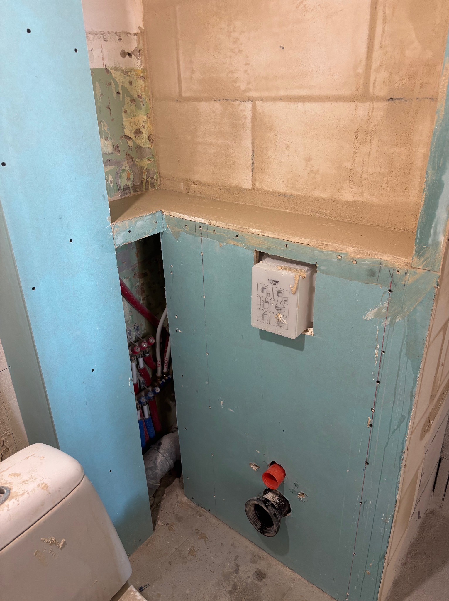 Narożnik łazienki w trakcie obudowy płytami GK, widoczne instalacje wod-kan, stelaż podtynkowy WC i fragment muszli. Surowy stan, przygotowanie do wykończenia.