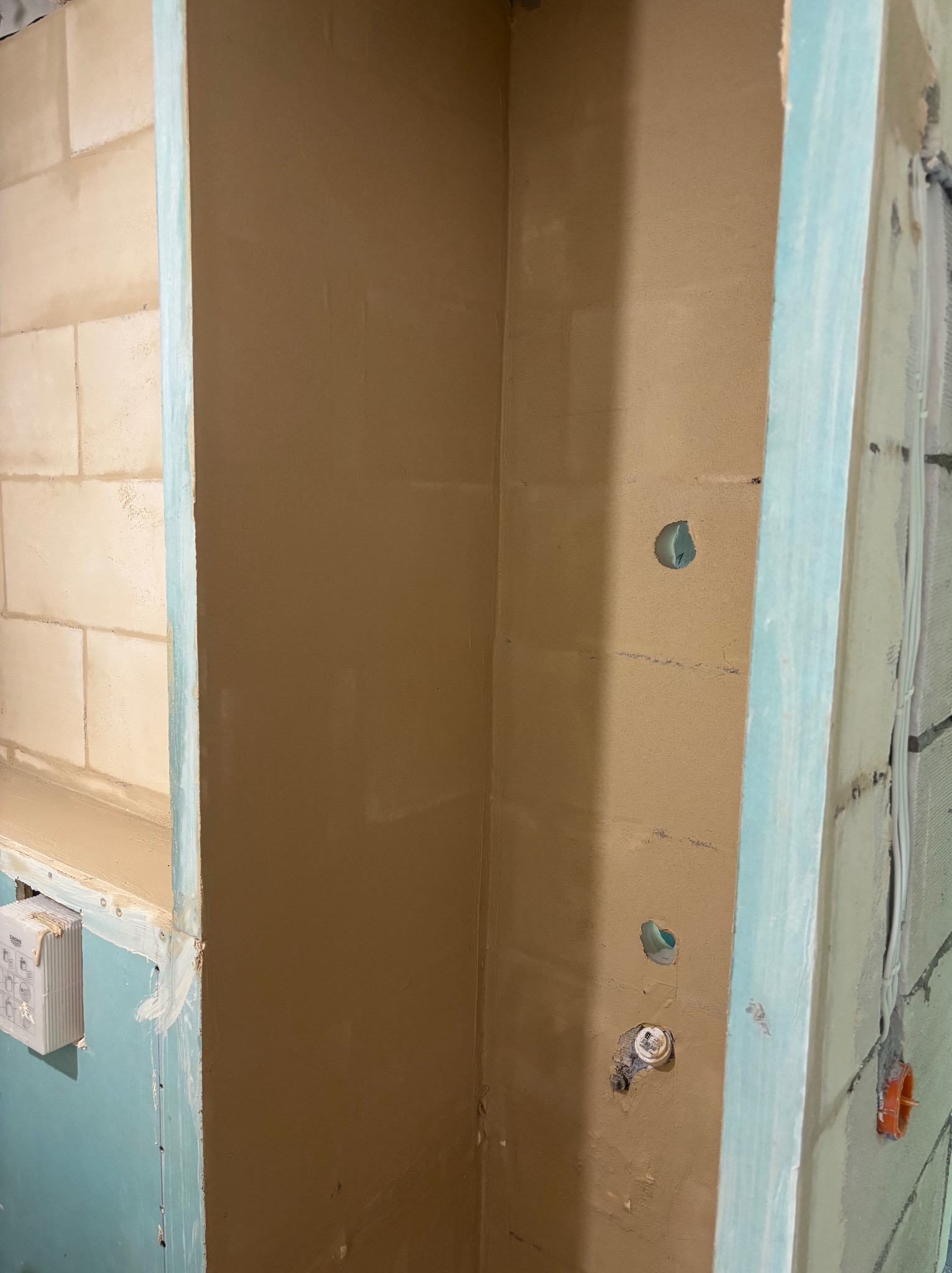 Ściana w trakcie szpachlowania z widocznymi otworami na instalacje, obudowana płytą gipsową z niebieskimi krawędziami. Fragment ściany z cegieł w tle.