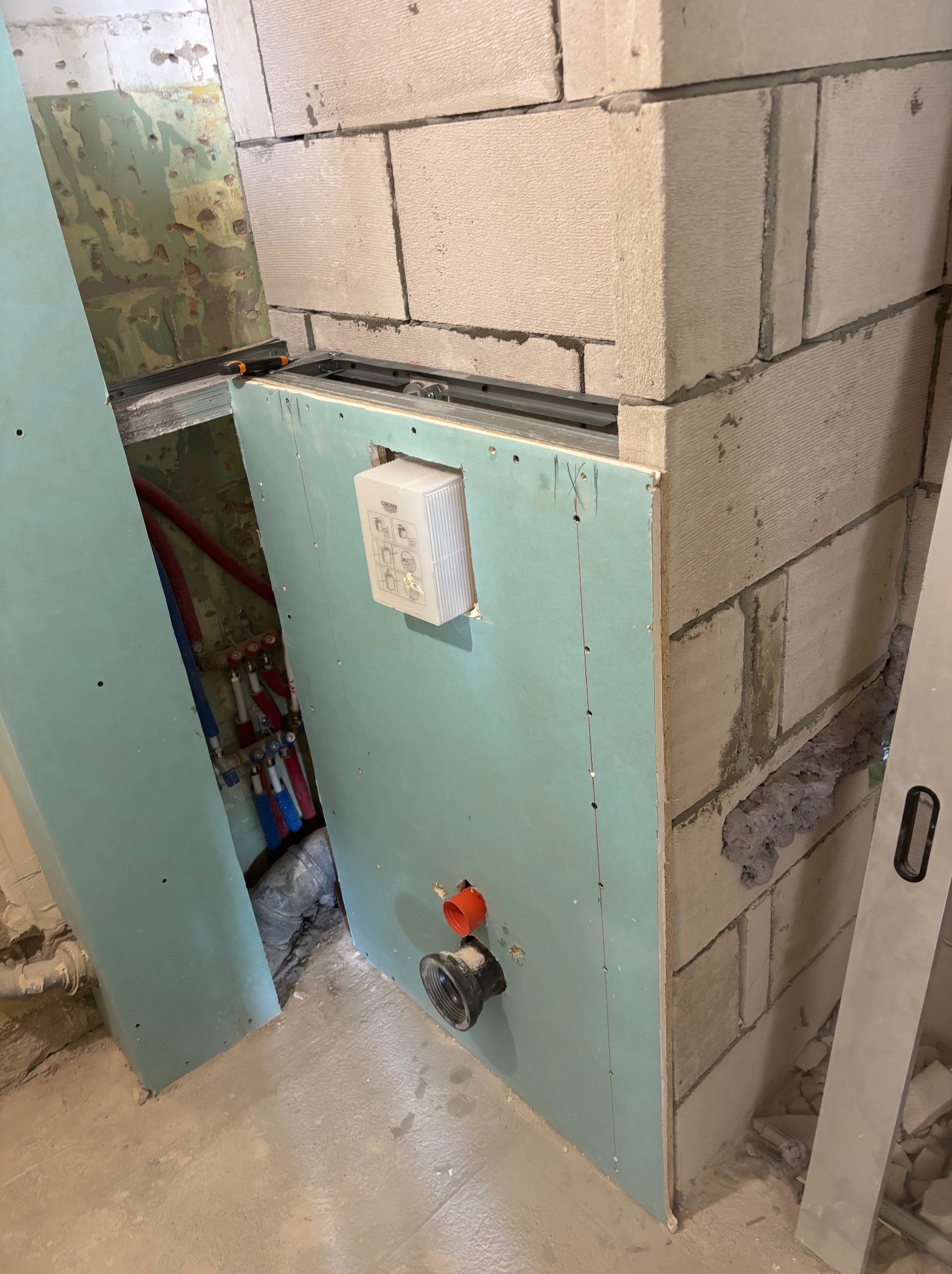 Ścianka działowa z płyty GK w trakcie montażu w łazience, widoczne instalacje wodne i kanalizacyjne. Surowy stan, bloczki betonowe, poziomica oparta o ścianę.