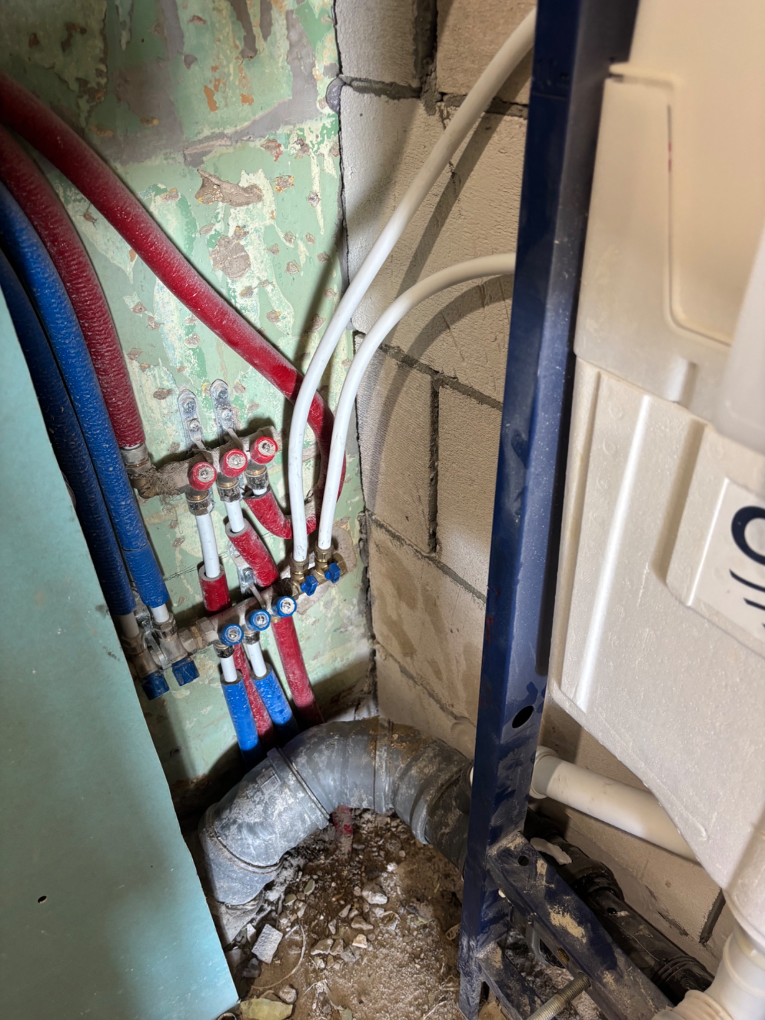Instalacja wodno-kanalizacyjna w trakcie montażu w łazience. Widoczne rury, zawory, stelaż podtynkowy i fragment ściany z widocznymi śladami starej farby.