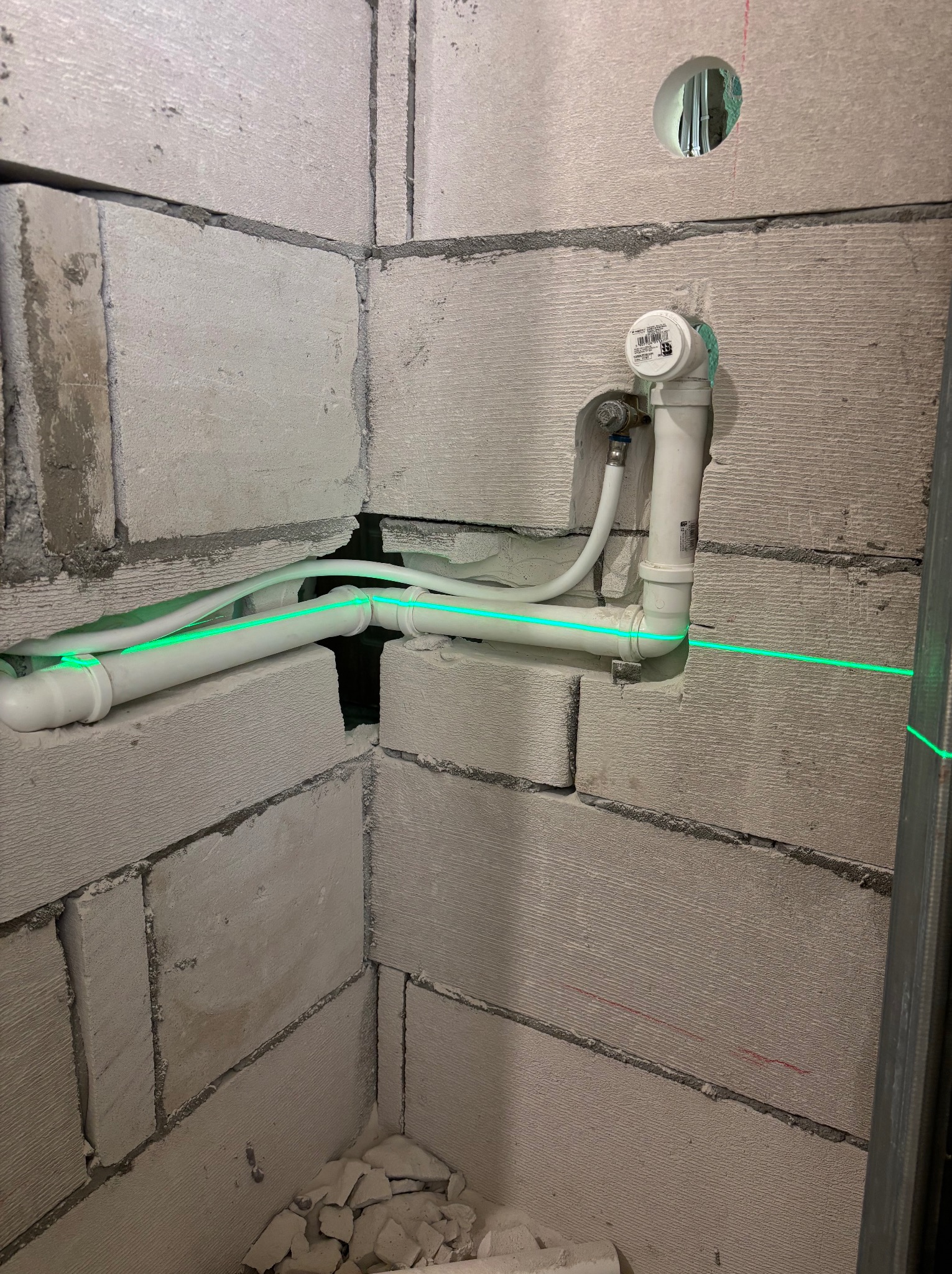 Narożnik łazienki w trakcie prac: białe rury instalacji wodnej, poziomica laserowa, ściany z szarego betonu komórkowego. Widoczne otwory i fragment metalowego profilu.