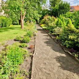 Ścieżka w ogrodzie z nowo posadzonymi roślinami, równo przycięty trawnik i ozdobne krzewy. Widok na dom w tle.