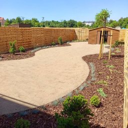 ZIELONY PATROL - Ogród z drewnianym płotem, ścieżką z piasku i rabatami wyłożonymi korą. Widoczny mały domek ogrodowy i świeżo posadzone drzewka.