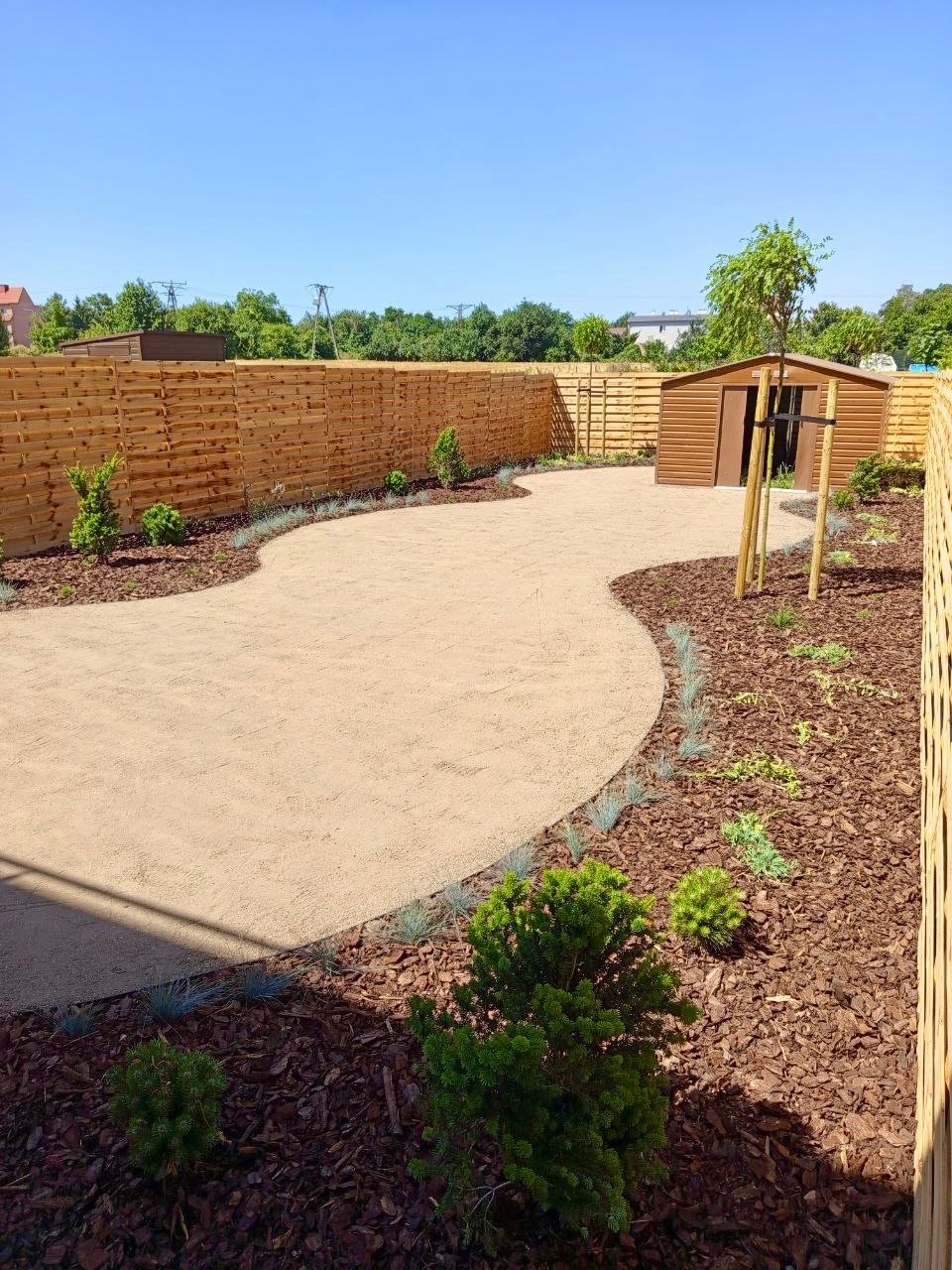 Ogród z drewnianym płotem, ścieżką z piasku i rabatami wyłożonymi korą. Widoczny mały domek ogrodowy i świeżo posadzone drzewka.