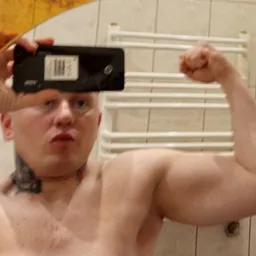 Mężczyzna robi selfie w lustrze, napinając biceps. Widoczny tatuaż na szyi, telefon w dłoni i fragment łazienki w tle. Oświetlenie neutralne, kadr centralny.