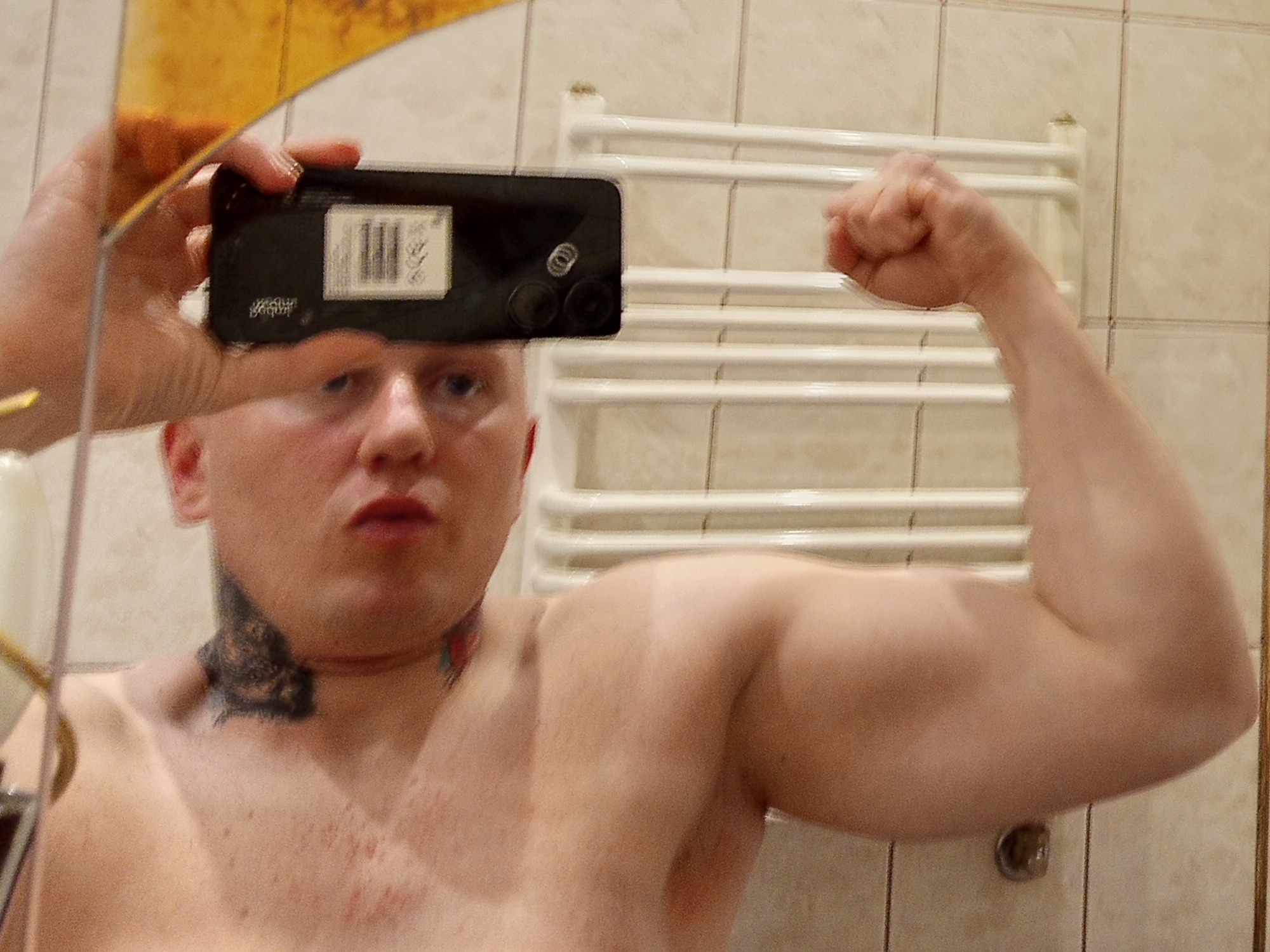 Mężczyzna robi selfie w lustrze, napinając biceps. Widoczny tatuaż na szyi, telefon w dłoni i fragment łazienki w tle. Oświetlenie neutralne, kadr centralny.