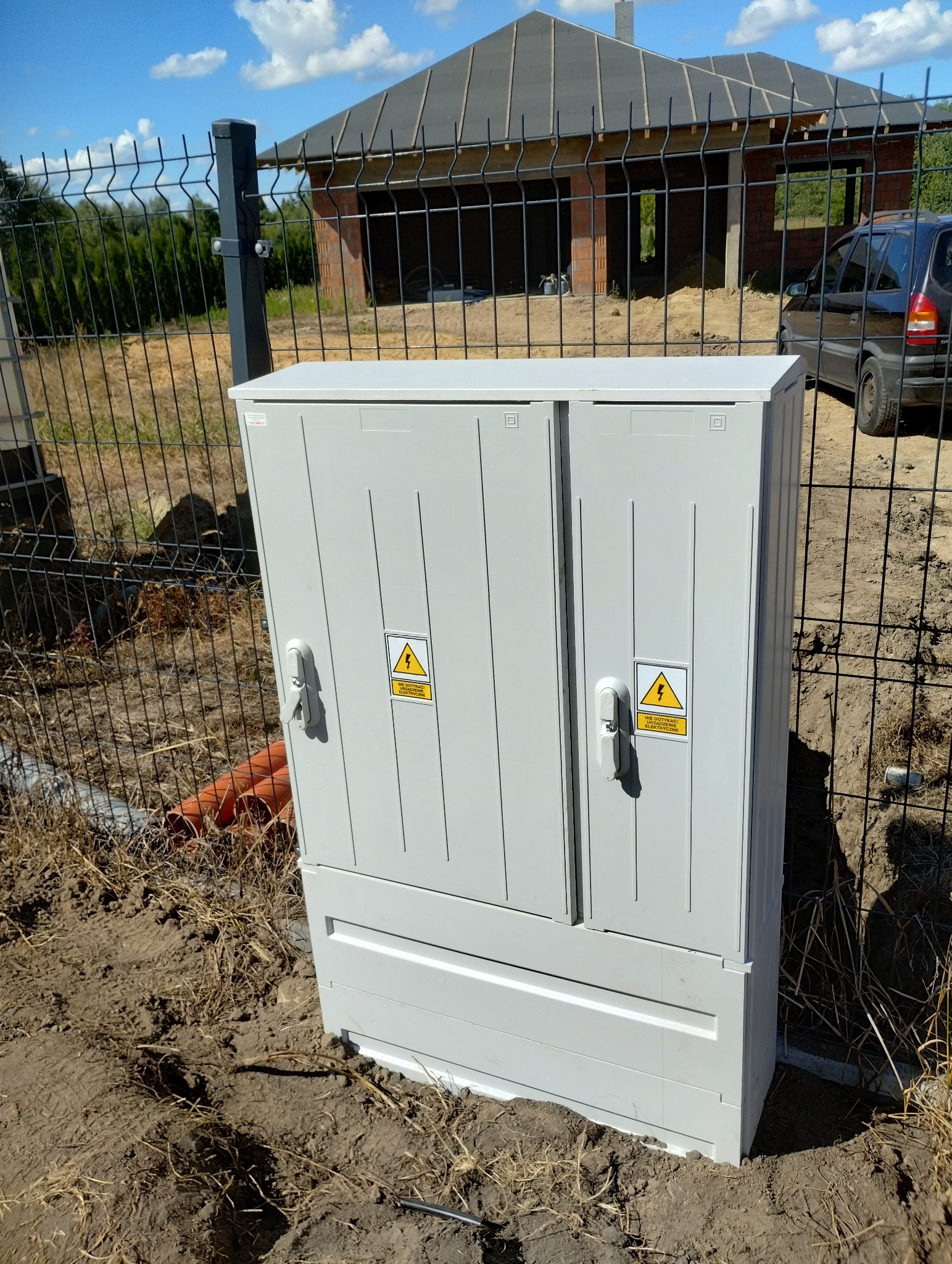 Szara skrzynka elektryczna z ostrzeżeniami, zamontowana przy ogrodzeniu na tle budowy domu i błękitnego nieba. Widoczne elementy instalacji.