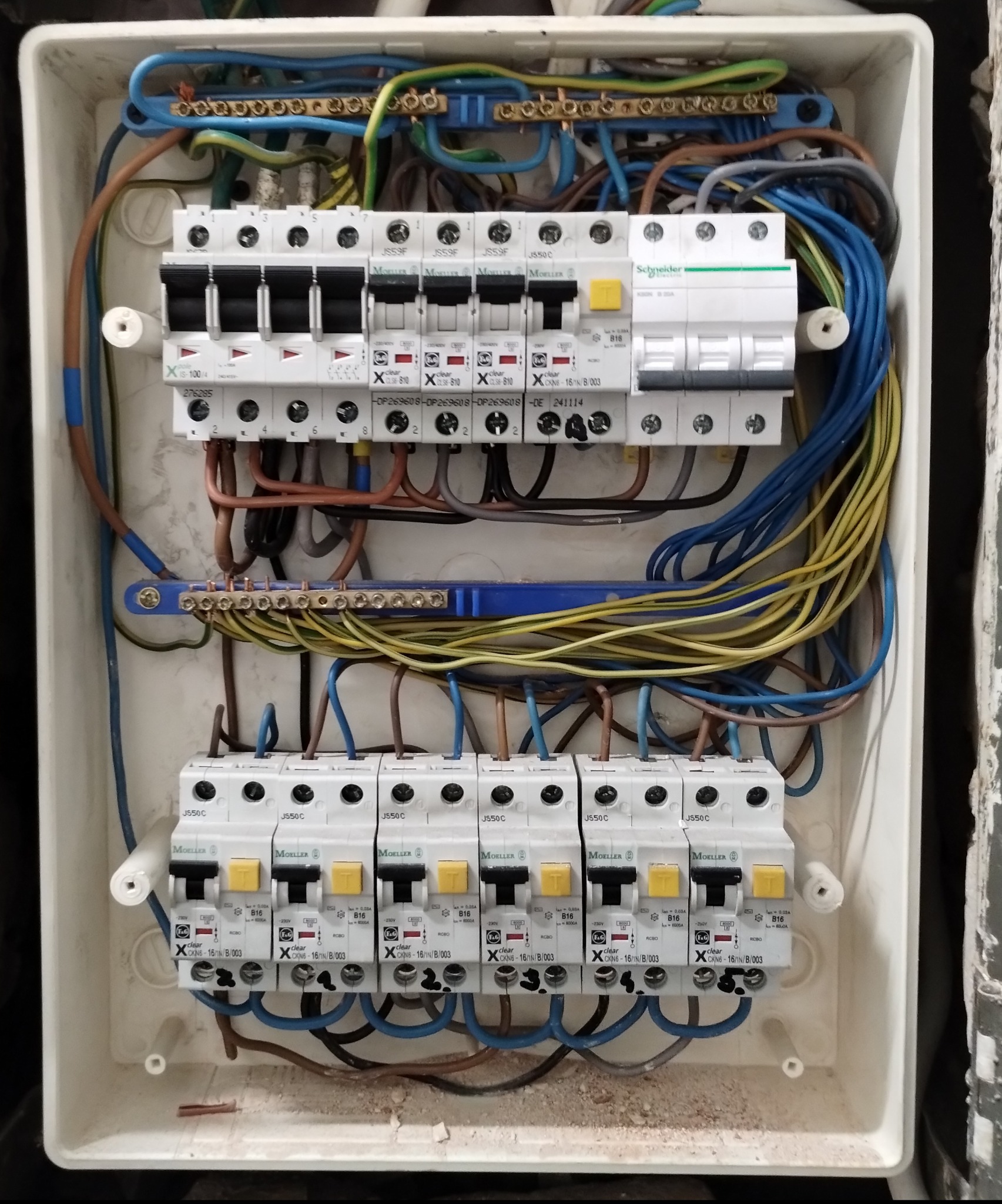 Otwarta skrzynka elektryczna z bezpiecznikami, widoczne przewody w kolorach: niebieskim, żółtym i brązowym. Instalacja w trakcie podłączania, detale montażu.