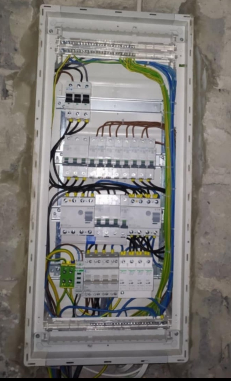 Kompletna, nowa rozdzielnia elektryczna w białej obudowie, z widocznymi zabezpieczeniami i okablowaniem, zamontowana na surowej, betonowej ścianie.