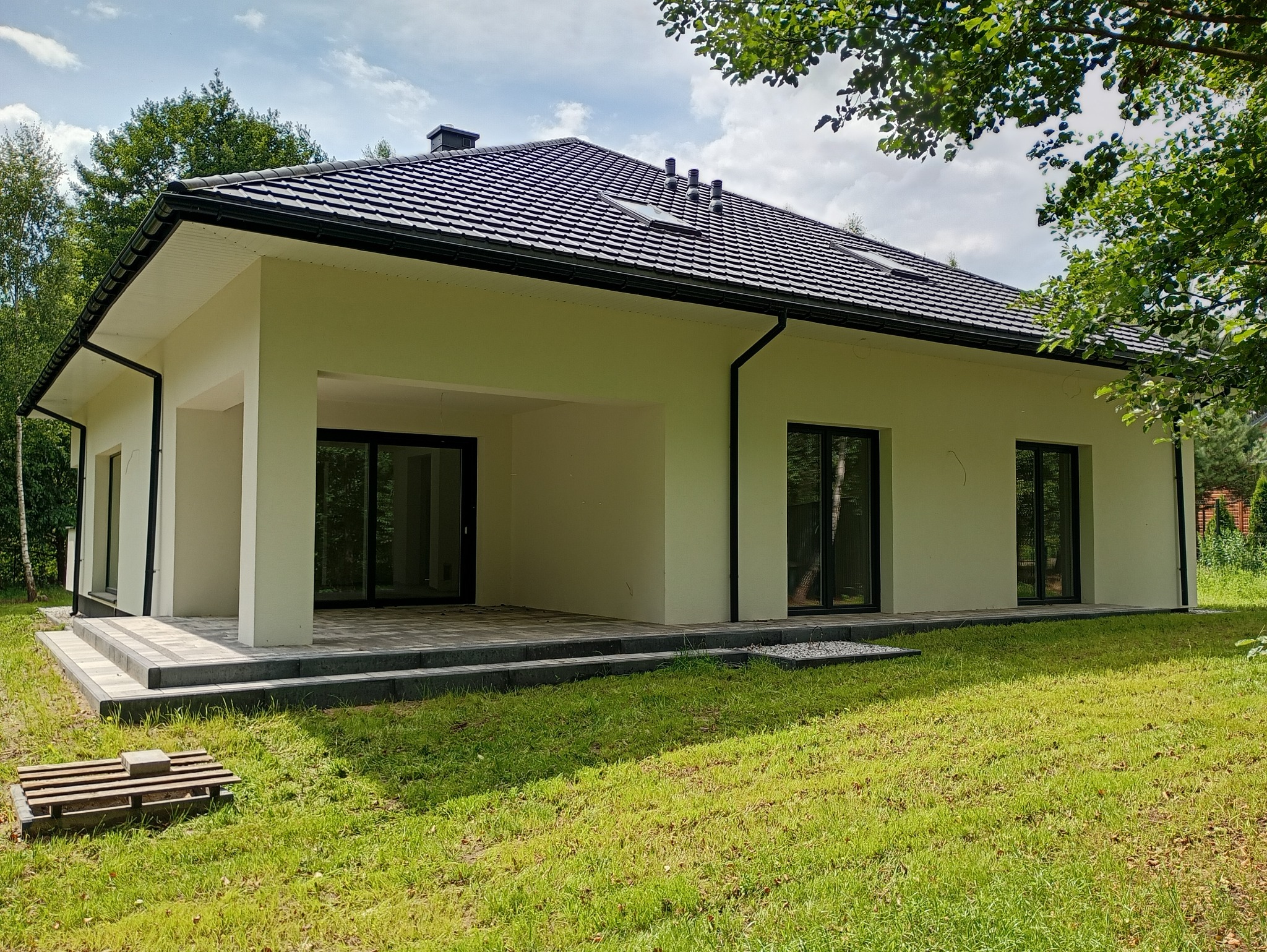 Parterowy dom z czarną dachówką i dużymi oknami, otoczony zielonym trawnikiem. Widoczne schody wejściowe i elementy wykończeniowe. Dom w stylu nowoczesnym.