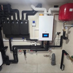 APE THERM SPÓŁKA Z OGRANICZONĄ ODPOWIEDZIALNOŚCIĄ - Złożona instalacja grzewcza z kotłem De Dietrich, zasobnikiem CWU i rozdzielaczami, rury w izolacji, manometry, zawory, całość na tle jasnych płytek.