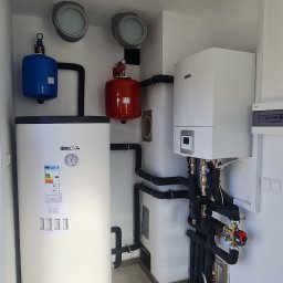 APE THERM SPÓŁKA Z OGRANICZONĄ ODPOWIEDZIALNOŚCIĄ - Biały bojler Kospel z czarnymi rurami i czerwonym zbiornikiem wyrównawczym w kotłowni. Widoczne elementy instalacji grzewczej i wentylacji.