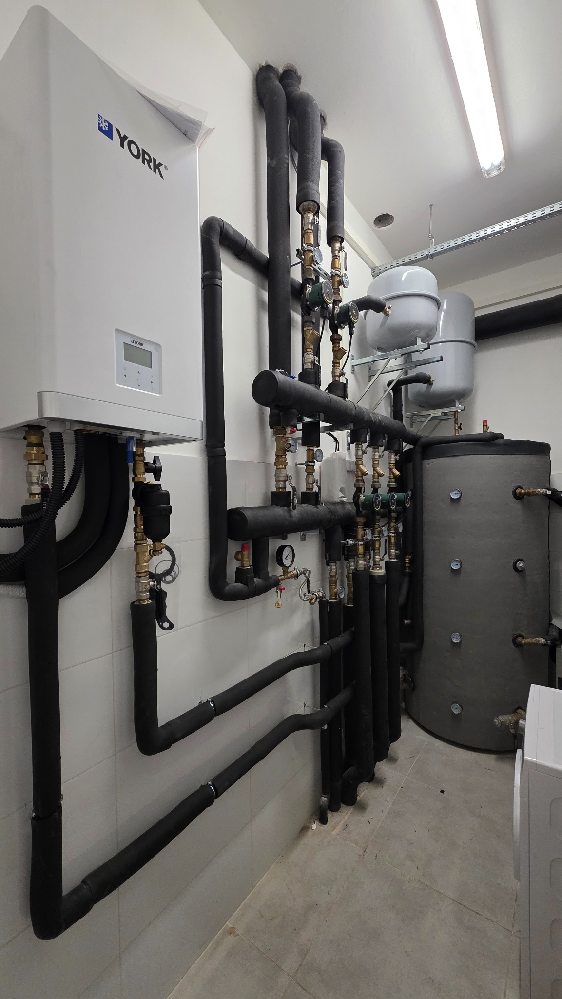 Instalacja grzewcza z piecem York, zbiornikiem i rurami w piwnicy z białymi kafelkami. Widoczne liczne zawory, manometry i czarne izolowane rury.
