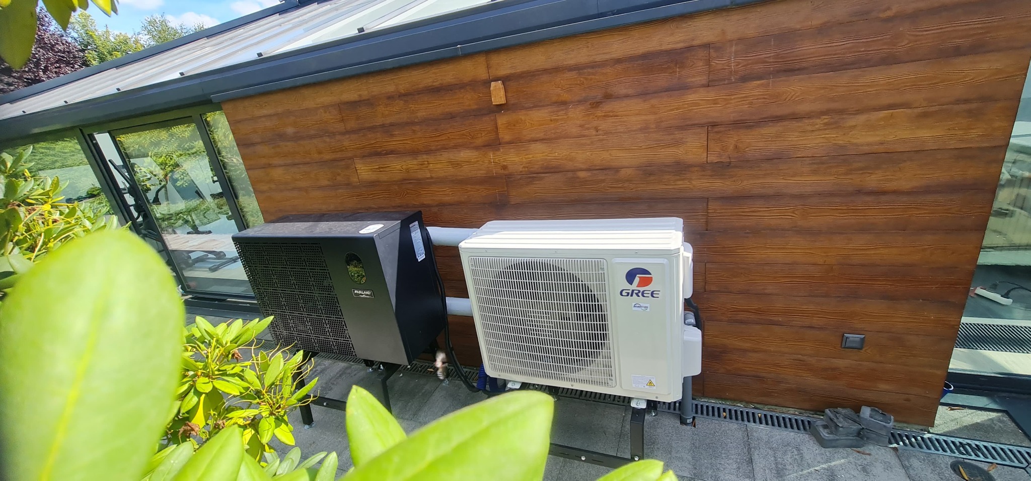 Dwa klimatyzatory (czarny i biały) zamontowane na zewnątrz budynku z drewnianą elewacją, widoczne zza zielonych liści. Jeden z klimatyzatorów marki GREE.