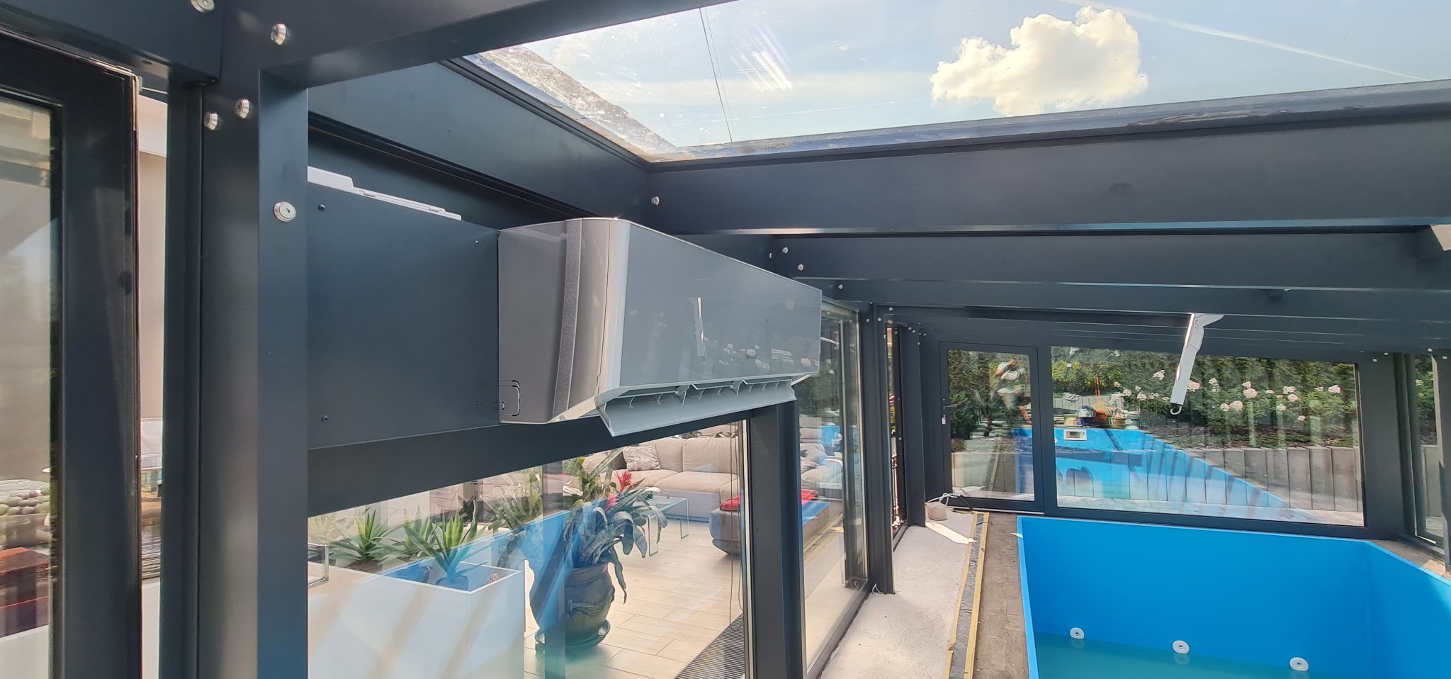 Szary klimatyzator zamontowany w nowoczesnej, przeszklonej oranżerii z basenem. Widok na błękitne niebo przez szklany dach. Minimalistyczny design.