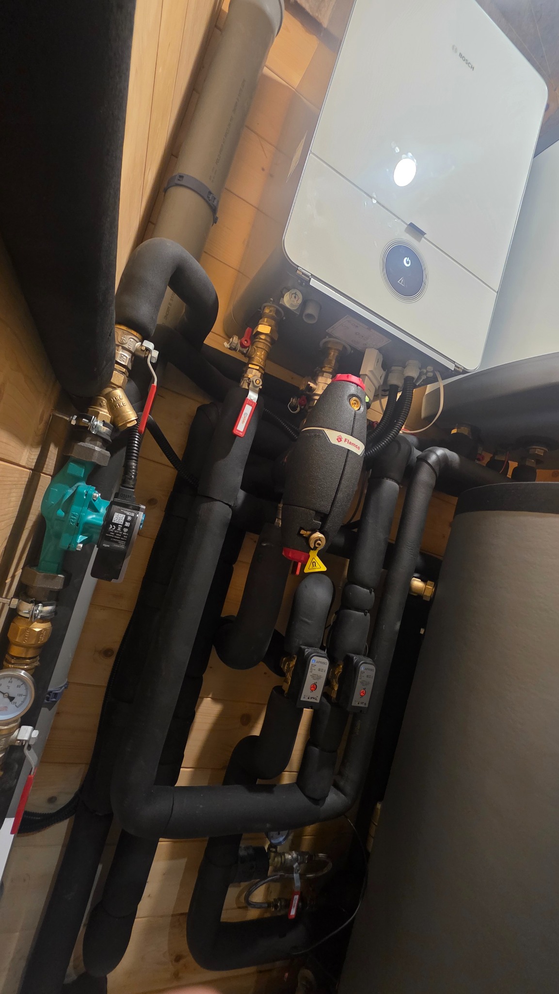 Nowoczesny piec gazowy Bosch z podłączeniami hydraulicznymi i czarnymi, izolowanymi rurami na tle drewnianej ściany. Widoczny zawór Flamco i manometr.