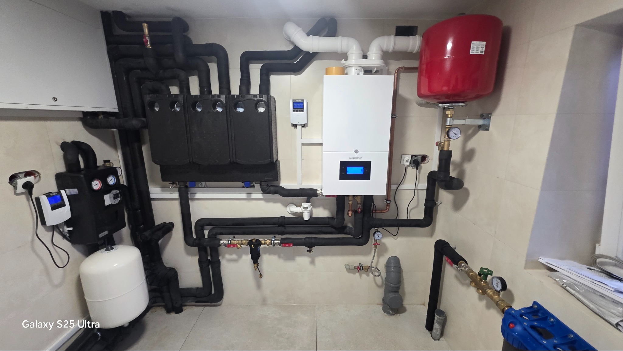 Złożona instalacja grzewcza z kotłem De Dietrich, zasobnikiem CWU i rozdzielaczami, rury w izolacji, manometry, zawory, całość na tle jasnych płytek.