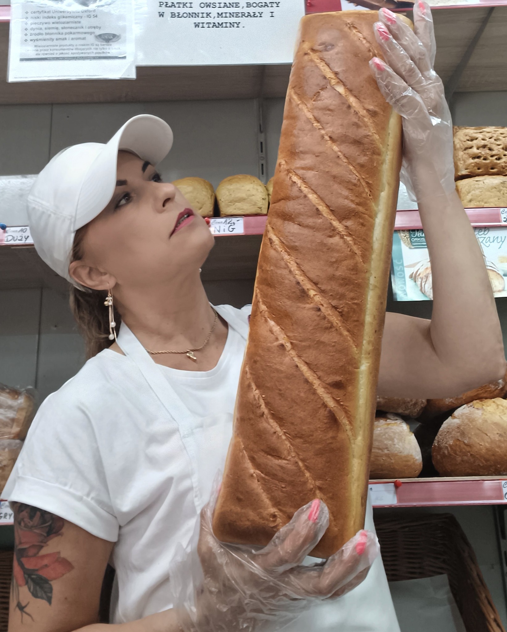 Kobieta w czapce i rękawiczkach prezentuje duży bochen chleba na tle półek z pieczywem w piekarni. Widoczne detale: tatuaż na ramieniu, biżuteria.