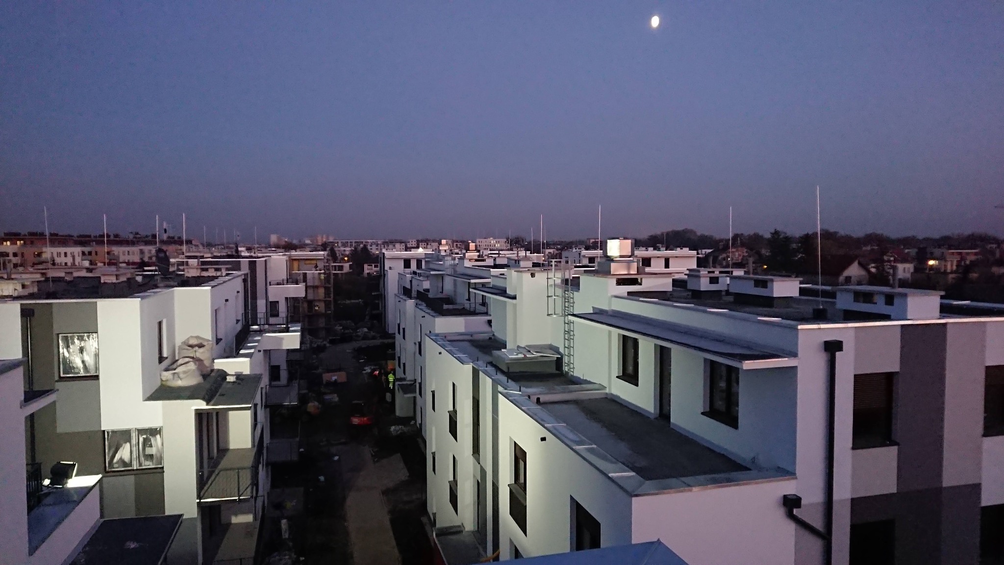 Osiedle nowych, wielopiętrowych domów z płaskimi dachami o zmierzchu, widziane z góry. W tle widoczne inne budynki i księżyc.