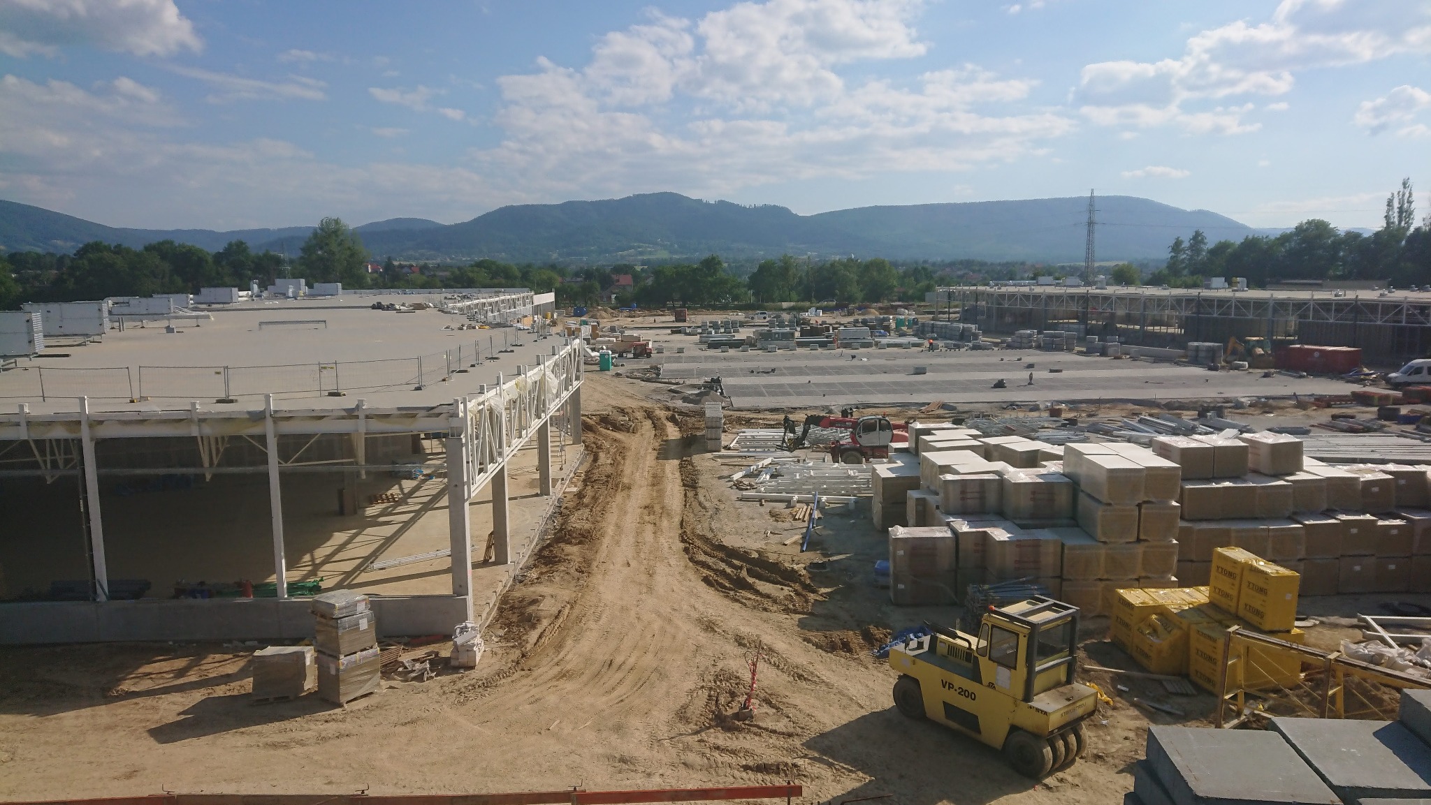 Szeroki kadr placu budowy w trakcie realizacji z żółtym walcem drogowym VP-200 na pierwszym planie i zarysem gór w tle.