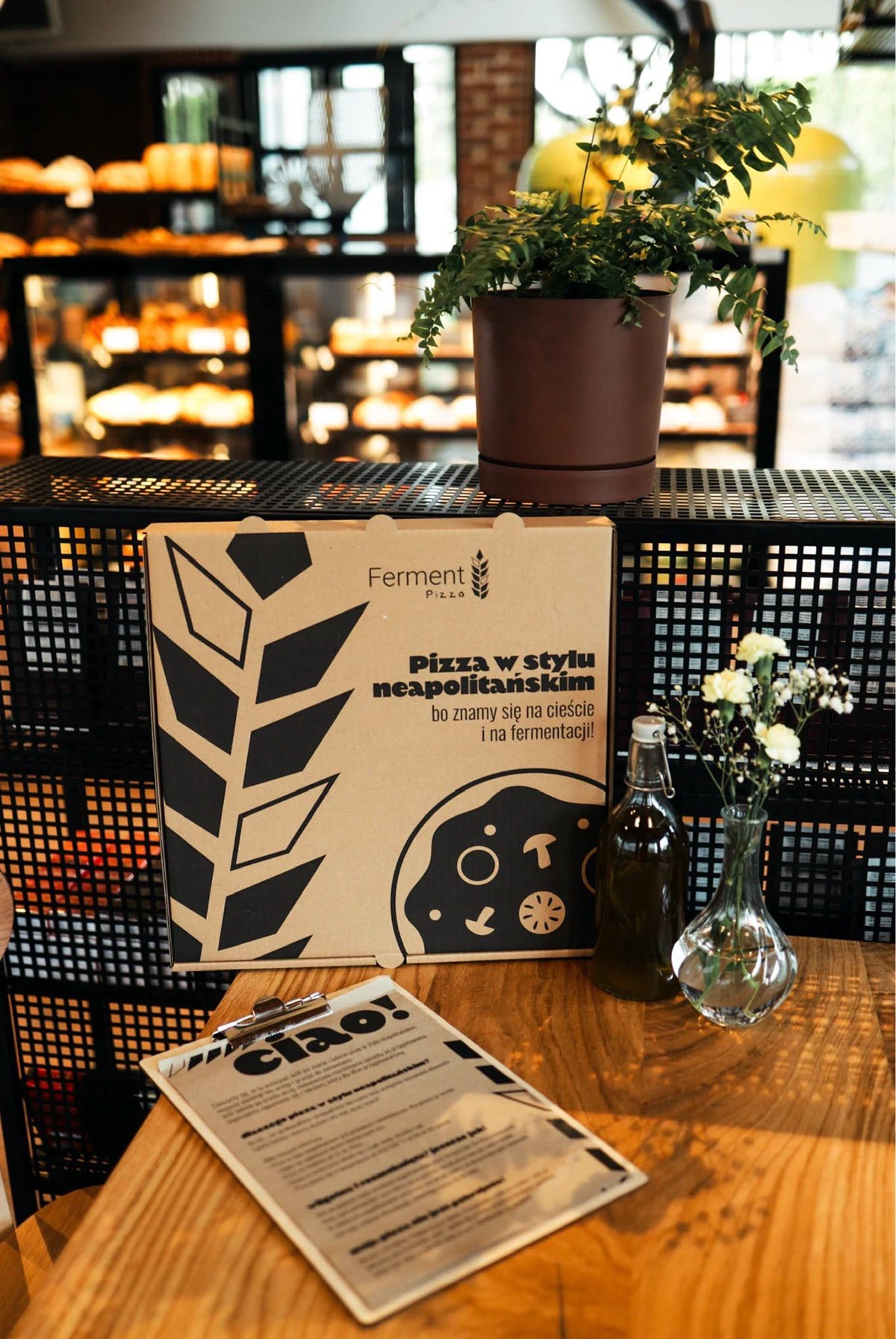 Karton pizzy 'Ferment Pizza' w stylu neapolitańskim, butelka oliwy i wazonik z kwiatami na drewnianym stoliku, w tle półki z pieczywem i roślina doniczkowa.