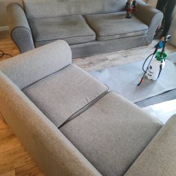 Lux House Pasja Sprzątania - Dwuosobowa szara kanapa z ciemnymi obwódkami poddawana czyszczeniu parowemu, obok stoi narożnik. Widoczny sprzęt do czyszczenia i osoba wykonująca usługę.