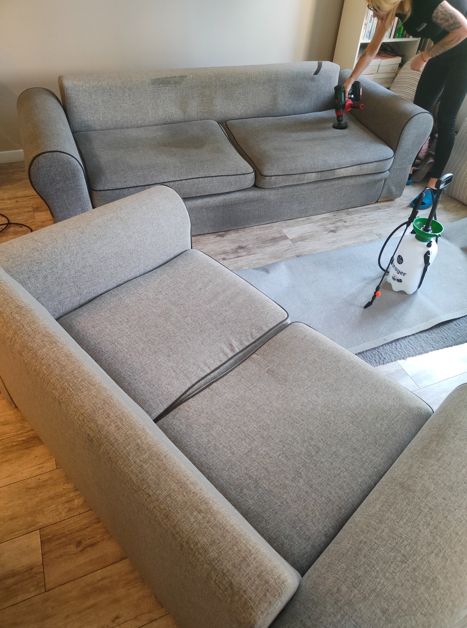 Dwuosobowa szara kanapa z ciemnymi obwódkami poddawana czyszczeniu parowemu, obok stoi narożnik. Widoczny sprzęt do czyszczenia i osoba wykonująca usługę.