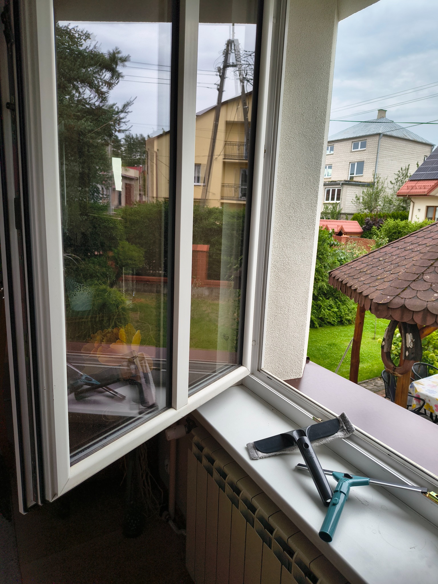 Otwarte okno z widokiem na zieleń i budynki. Na parapecie leżą narzędzia do mycia okien: ściągaczka z gumą i myjka z mikrofibrą.