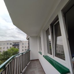Projekt Home - Odnowiony balkon z szarą terakotą, białą balustradą i zielonymi parapetami. Widok na budynek mieszkalny w tle, pochmurne niebo.