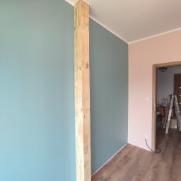 Projekt Home - Pomieszczenie po malowaniu ścian na dwa kolory (turkusowy i różowy) z drewnianą kolumną. Widoczna podłoga i drabina w tle.