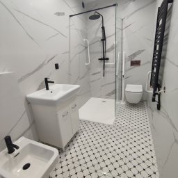 Projekt Home - Elegancka, wykończona łazienka w bieli i czerni. Kabina prysznicowa ze szklaną ścianką, umywalka z szafką, bidet i grzejnik drabinkowy. Płytki podłogowe w geometryczny wzór.