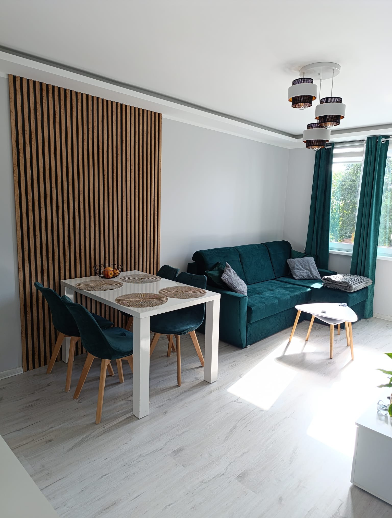 Nowoczesny salon z drewnianą ścianką działową, turkusową sofą i białym stołem z krzesłami. Jasne panele podłogowe i okno z zasłonami dopełniają wystrój.