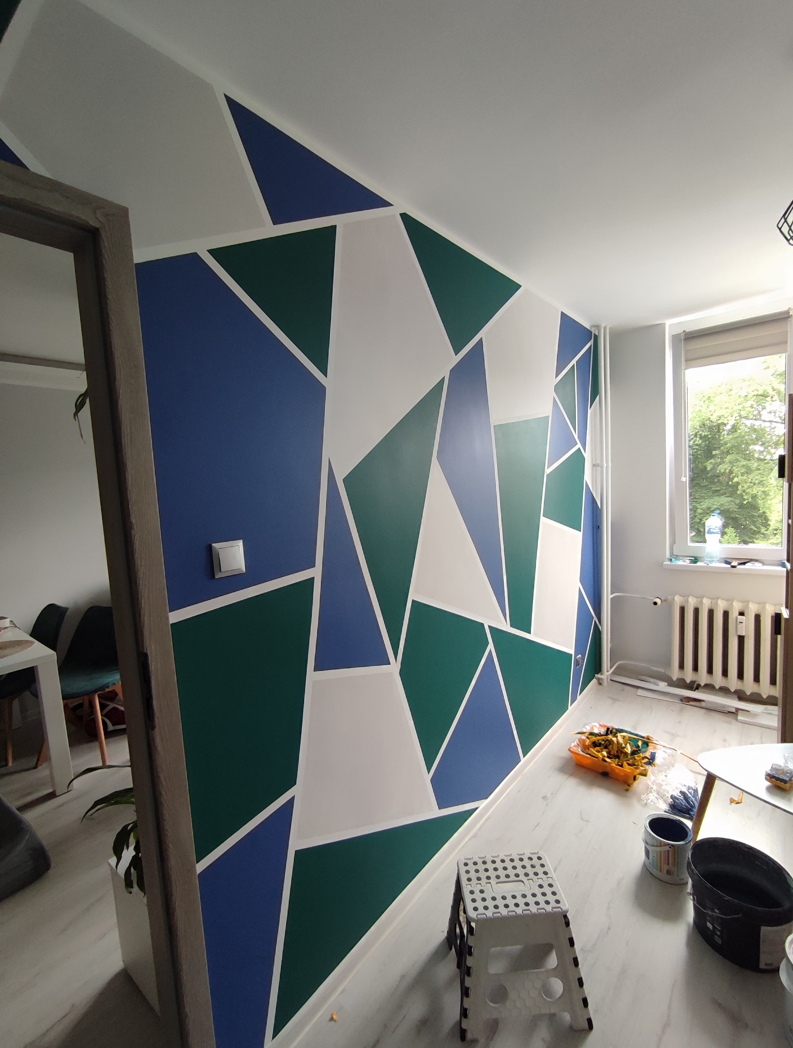 Pomysłowo pomalowana ściana w geometryczne wzory w odcieniach bieli, zieleni i niebieskiego. Widoczne narzędzia malarskie i drabinka.