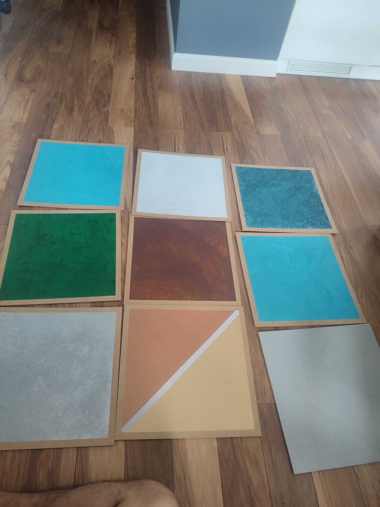 Paleta kolorów farb w Koszalinie. Kwadratowe próbki w odcieniach zieleni, brązu, błękitu i szarości ułożone na drewnianej podłodze.