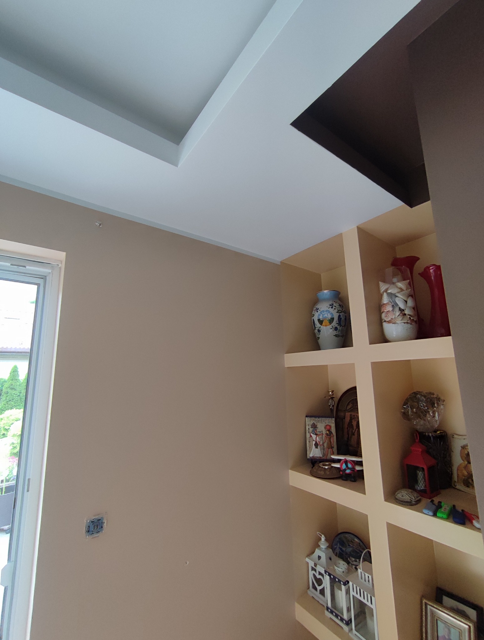 Beżowa ściana z gładzią, biały sufit podwieszany z brązowym wnęką, półka z dekoracjami. Widoczny fragment okna.