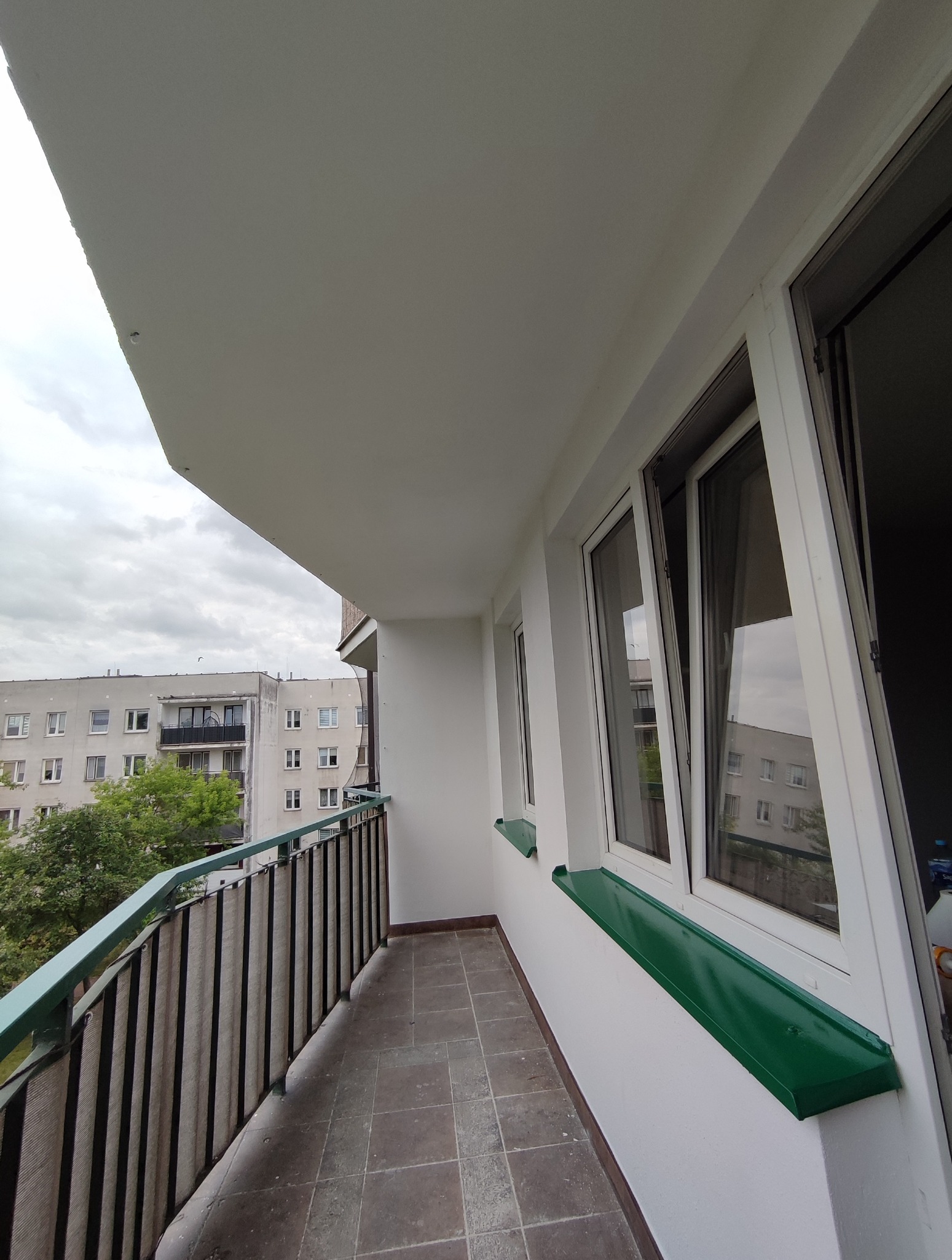 Odnowiony balkon z szarą terakotą, białą balustradą i zielonymi parapetami. Widok na budynek mieszkalny w tle, pochmurne niebo.