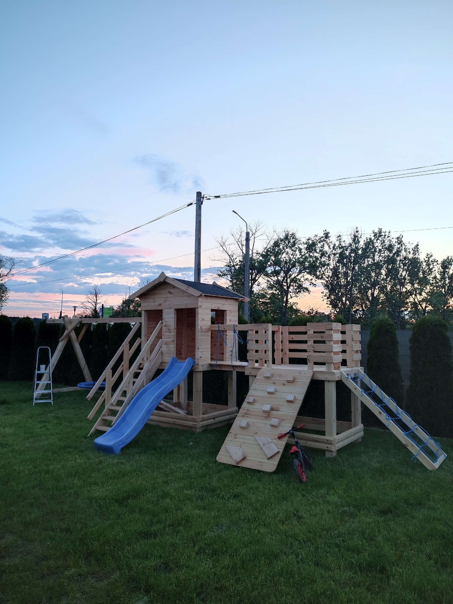 Drewniany plac zabaw z domkiem, zjeżdżalnią i ścianką wspinaczkową na zielonym trawniku, oświetlony wieczornym światłem. W tle widoczne drzewa i niebo.