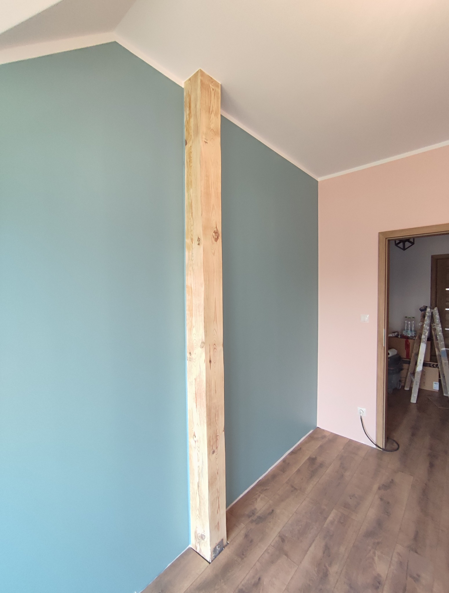 Pomieszczenie po malowaniu ścian na dwa kolory (turkusowy i różowy) z drewnianą kolumną. Widoczna podłoga i drabina w tle.