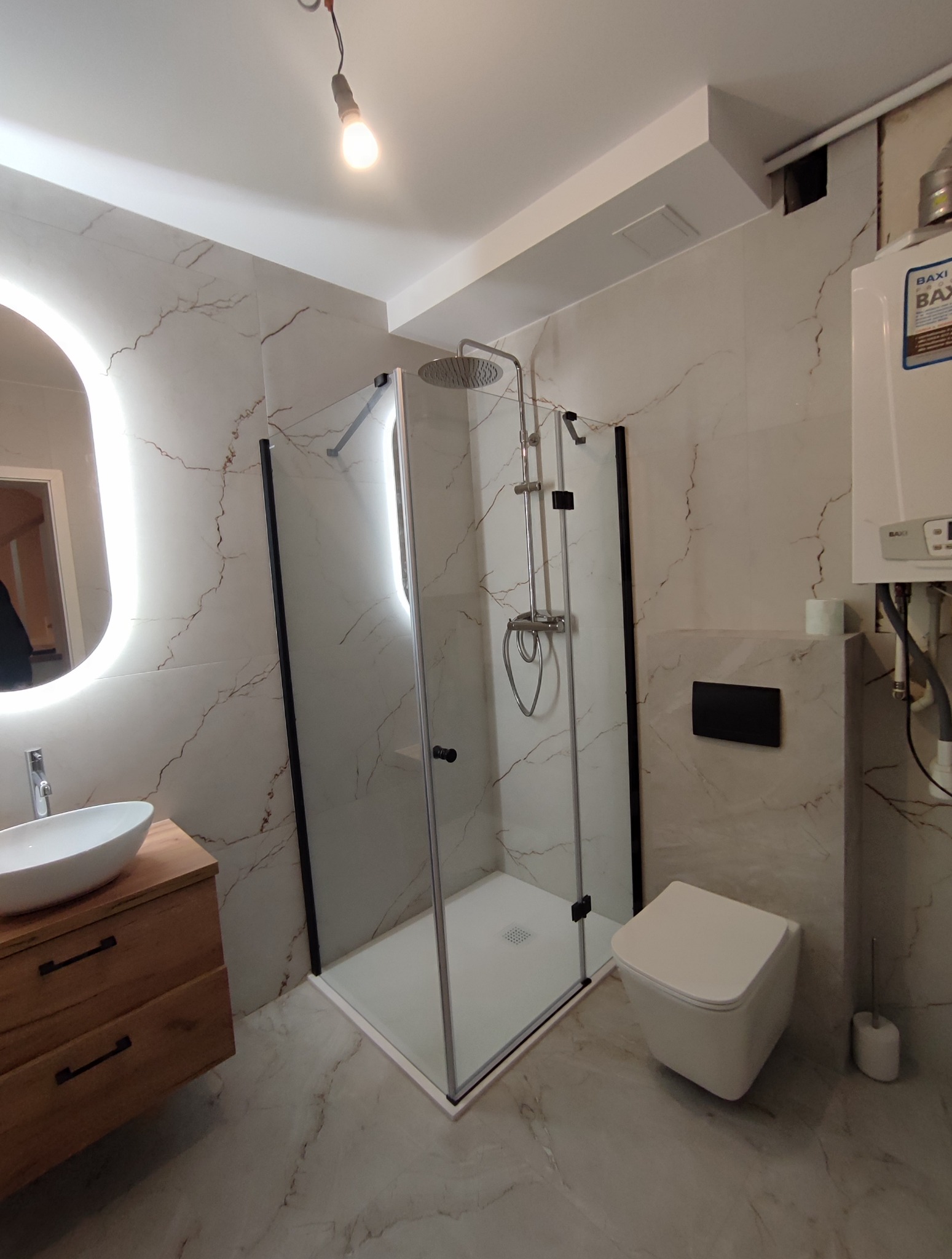 Nowoczesna łazienka z kabiną prysznicową typu walk-in, umywalką na drewnianej szafce i podświetlanym lustrem. Ściany wykończone marmurkową płytką.