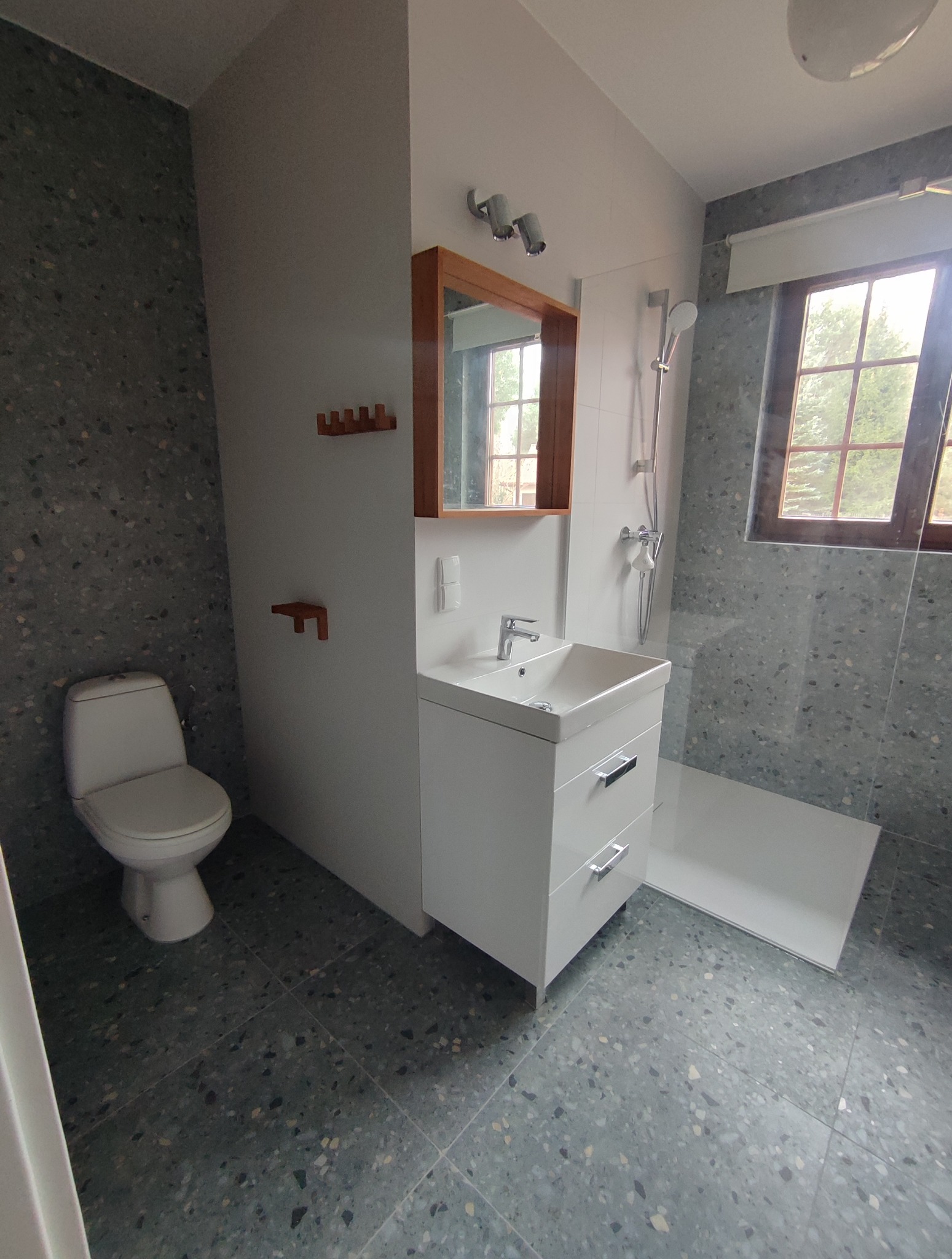 Nowoczesna łazienka z szarą mozaiką, białą szafką z umywalką i lustrem w drewnianej ramie. Widoczny fragment kabiny prysznicowej i toaleta.