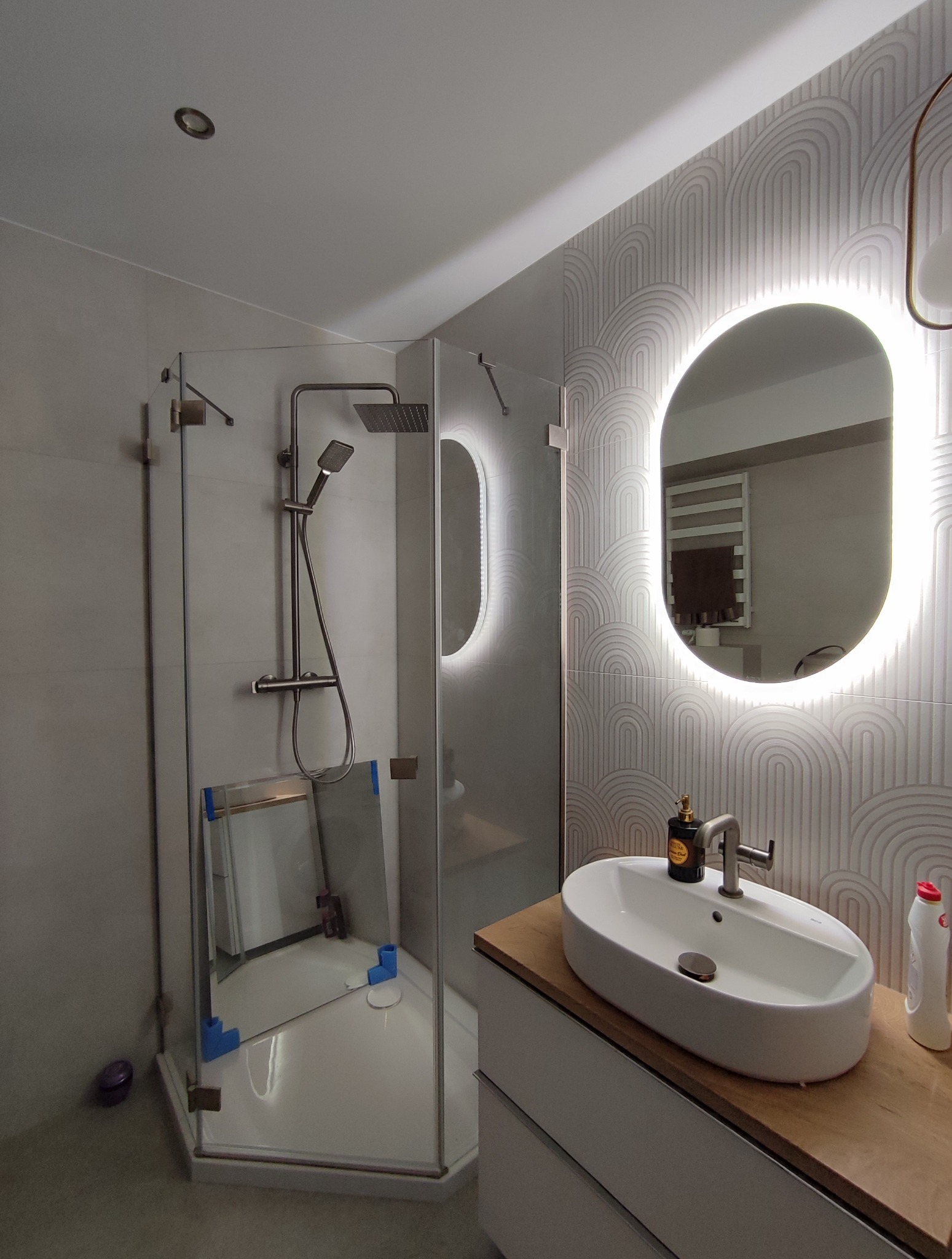 Nowoczesna łazienka po remoncie z kabiną prysznicową typu walk-in, umywalką nablatową i podświetlanym lustrem. Minimalistyczny design z geometrycznym wzorem na ścianie.