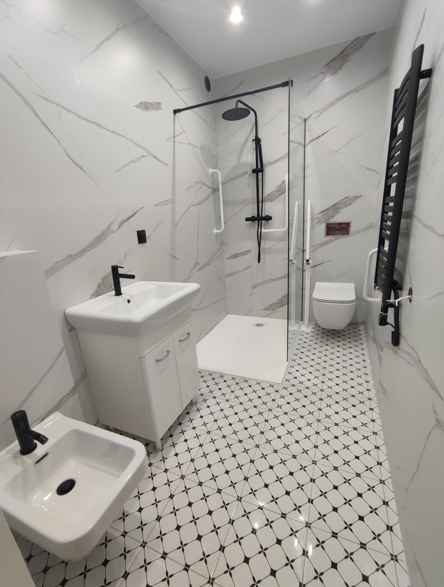 Elegancka, wykończona łazienka w bieli i czerni. Kabina prysznicowa ze szklaną ścianką, umywalka z szafką, bidet i grzejnik drabinkowy. Płytki podłogowe w geometryczny wzór.