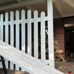 Białe, pomalowane ogrodzenie z desek, częściowo zasłonięte przez aluminiową drabinę. W tle widoczny budynek z cegły i ułożone drewno.