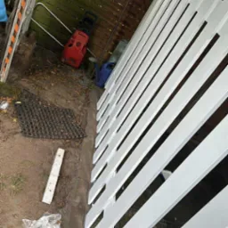 Świeżo pomalowane, białe ogrodzenie drewniane, widoczne z bliska, z elementami otoczenia jak drabina i myjka ciśnieniowa w tle. Ślady farby na podłożu.