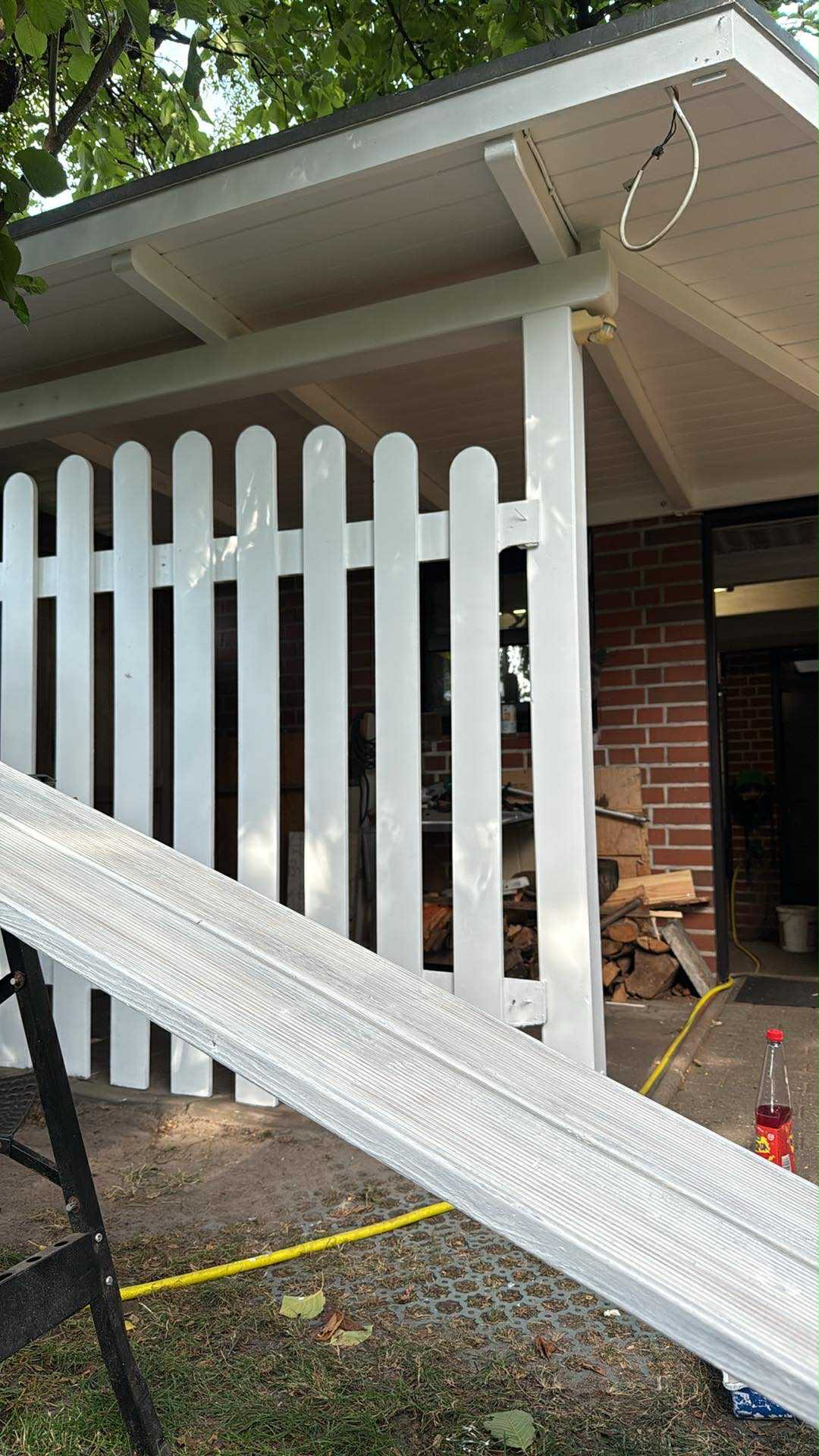 Białe, pomalowane ogrodzenie z desek, częściowo zasłonięte przez aluminiową drabinę. W tle widoczny budynek z cegły i ułożone drewno.