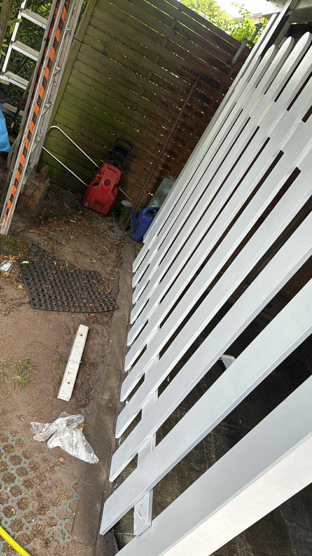 Świeżo pomalowane, białe ogrodzenie drewniane, widoczne z bliska, z elementami otoczenia jak drabina i myjka ciśnieniowa w tle. Ślady farby na podłożu.